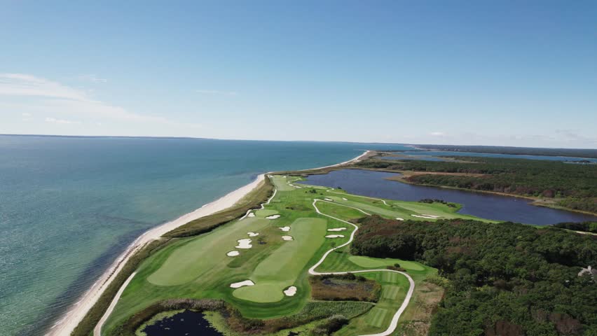 Aerial view of a lush green golf course, bordered by the deep blue ocean on one side and a dark lake surrounded by dense trees, Boston, Massachusetts, United States.