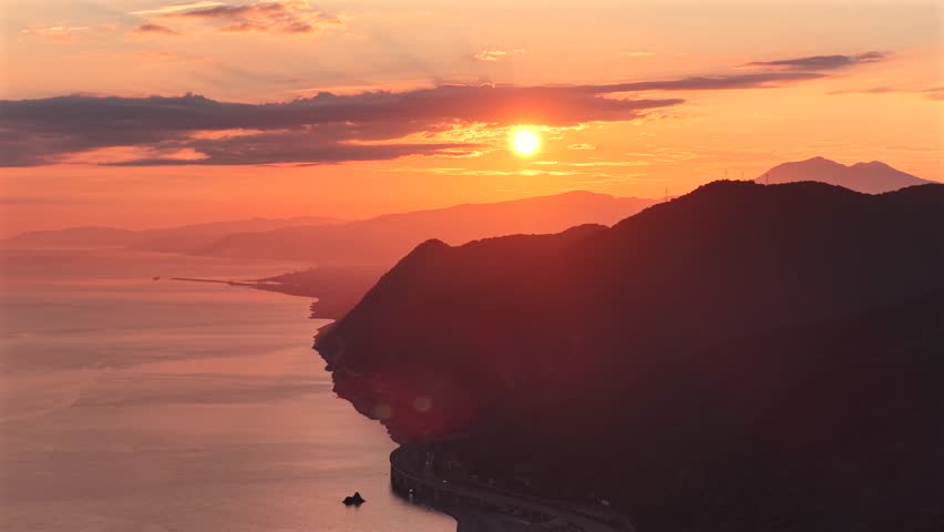 Aerial view of the sun setting over the sea, casting an orange glow on the mountains and coastline, Sendai, Miyagi, Japan.