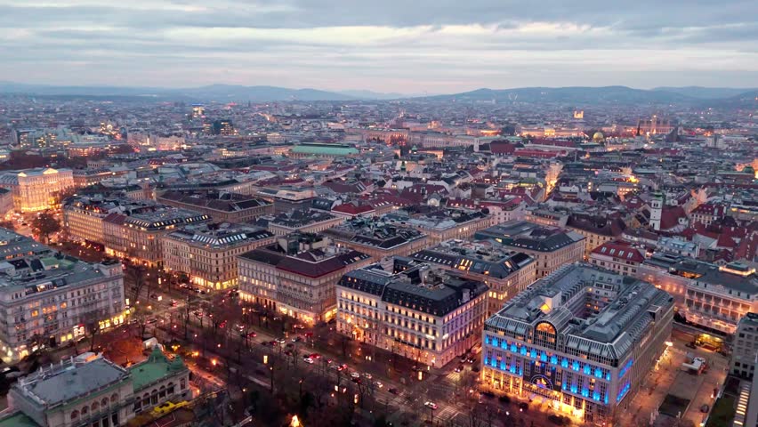 Aerial drone shot panning left over Vienna cityscape at night, showing illuminated streets, glowing traffic lines, dense urban buildings, and low cloudy skies reflecting city light across the skyline.
