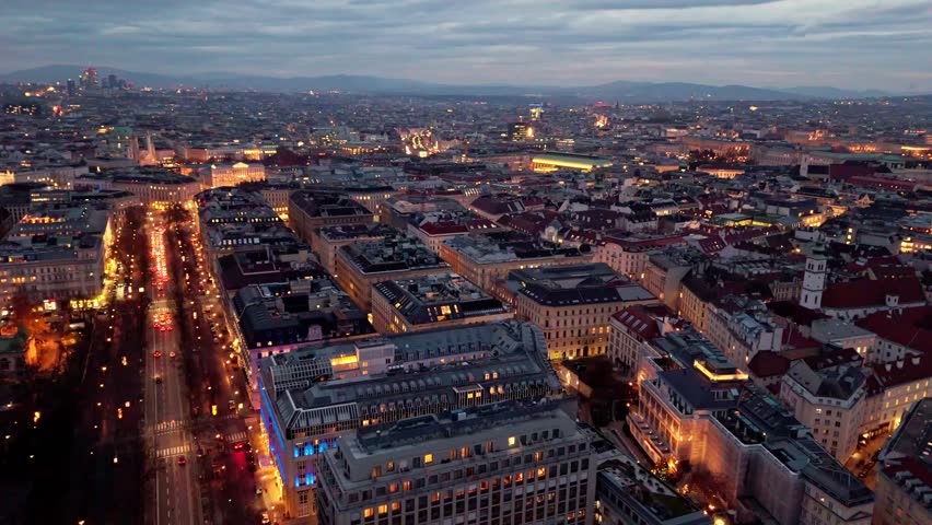 Aerial landscape drone shot panning left over Vienna cityscape at night, showing illuminated streets, dense urban buildings, winter atmosphere, glowing traffic lines, and city lights spreading