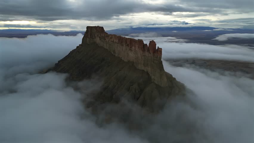 Aerial view of the majestic mountain, its peak piercing through a sea of swirling clouds, casting a dramatic silhouette against the muted horizon, Hanksville, Utah, United States.