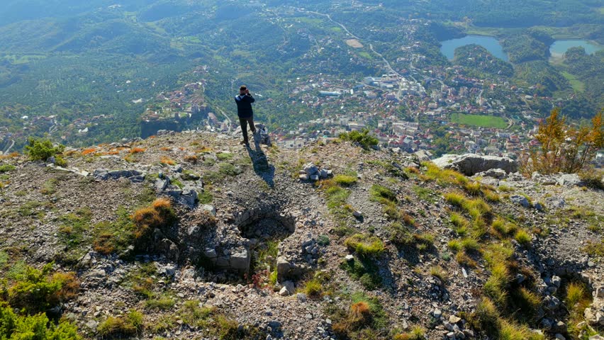 Aerial video of a photographer standing on rocky slopes of Krujë Mountain with the city and valley below in the distance.