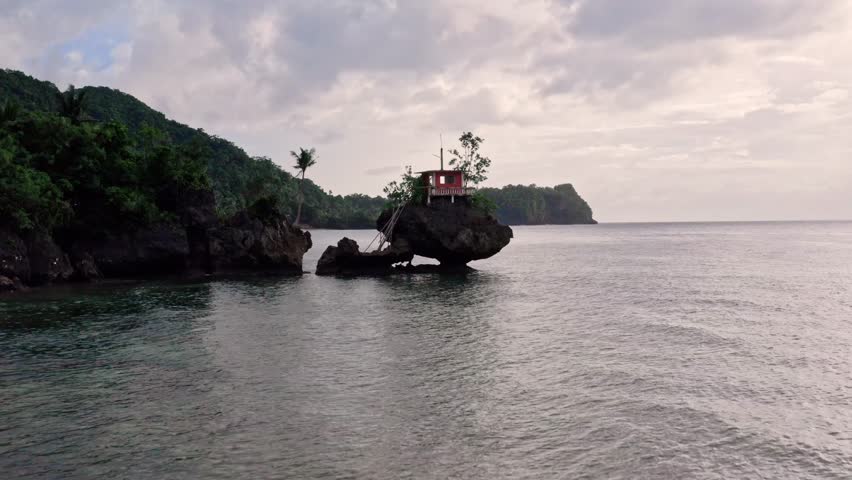 Aerial view of a red house perched atop a dark, rocky formation surrounded by calm waters contrasting with the lush green hills, Buruanga, Western Visayas, Philippines.