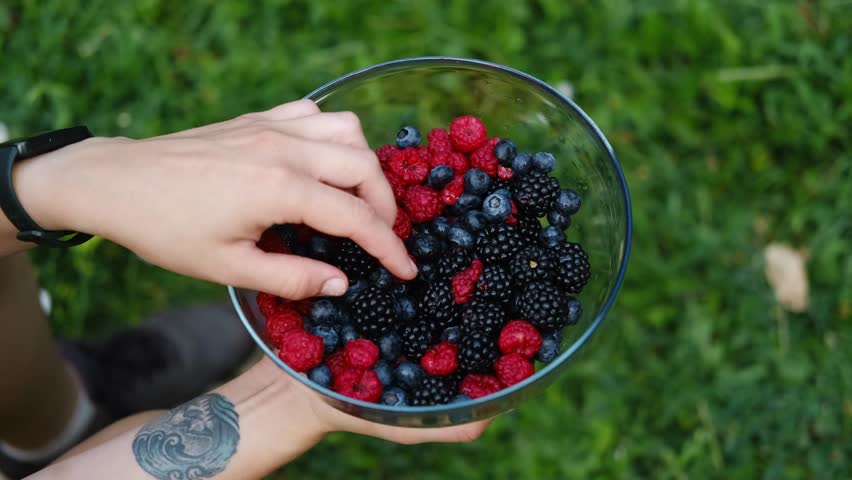 Close-up of woman hands holding a glass bowl filled with fresh raspberries, blueberries, and blackberries while picking one berry. Concept of healthy lifestyle, freshness, and summer nutrition