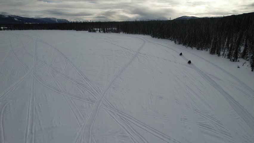 Tourists ride snowmobiles across the wide, frozen surface of Murray Lake in the Yukon wilderness, surrounded by boreal forest and snow-dappled mountains on a winter day