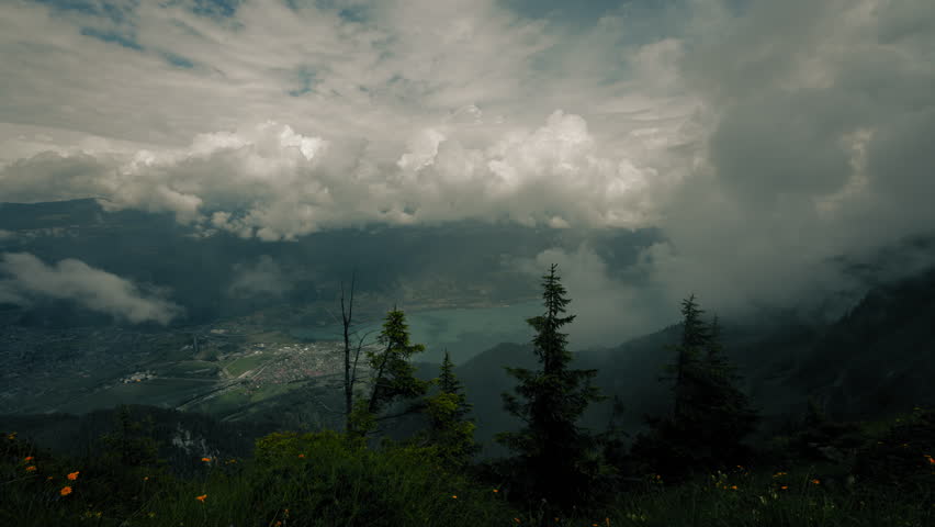Wide panoramic view from a high mountain ridge overlooking a Swiss alpine valley with a turquoise lake below, dramatic clouds rolling across the sky and mist drifting through evergreen forests
