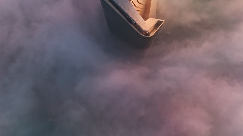Close-up drone aerial of a modern skyscraper rooftop cutting through dense clouds at sunrise, golden light on the facade with fog swirling over the hidden city below.