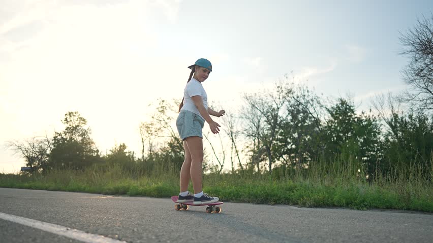 The girl confidently rides her skateboard down the road, learning how to control every move. The skateboard helps the girl feel more free, while the outdoor setting makes her ride exciting and joyful.