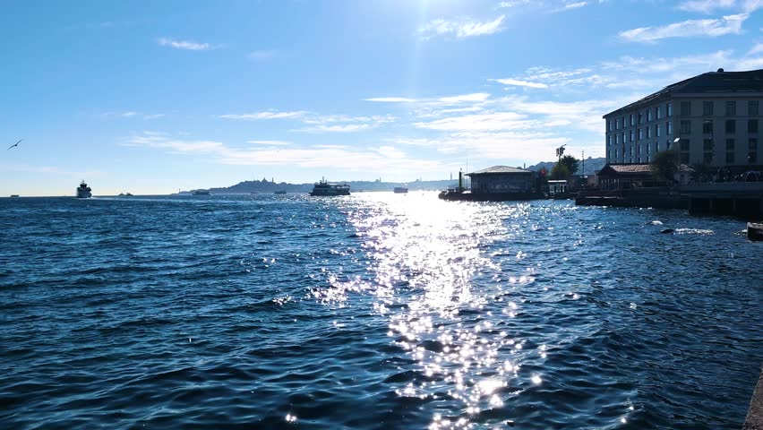Captivating wide shot of the Bosphorus Strait in Istanbul, Turkey, captured from the Besiktas shoreline. The video showcases the intense and mesmerizing shimmer of bright sunlight dancing on the deep blue wavy waters, creating a dazzling sparkling effect. In the background, the silhouette of the historical peninsula, including Topkapi Palace and Hagia Sophia, can be faintly seen under a clear blue sky with soft white clouds.

The scene features the Besiktas ferry terminal and adjacent historical stone buildings, reflecting the unique blend of Ottoman-era architecture and modern maritime life. Public ferries (Vapur) and private boats are seen navigating the busy waterway, which serves as a vital bridge between Europe and Asia. This high-quality footage captures the serene yet vibrant atmosphere of a sunny day in Istanbul, making it perfect for travel documentaries, tourism promotions, cultural projects, or cinematic transitions. The natural lighting emphasizes the texture of the sea and the grandeur of the city
