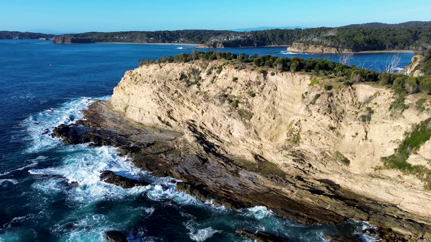 Drone aerial landscape with ocean waves crashing on rocky headland cliffs rugged terrain covered bushland trees overlooking Rosedale suburban town in Malua Bay South Coast Australia nature tourism