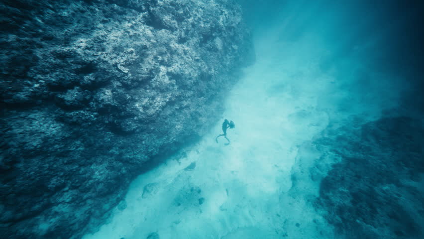 Diver ascends slowly beside massive reef rock as sandy seabed fades into blue light below