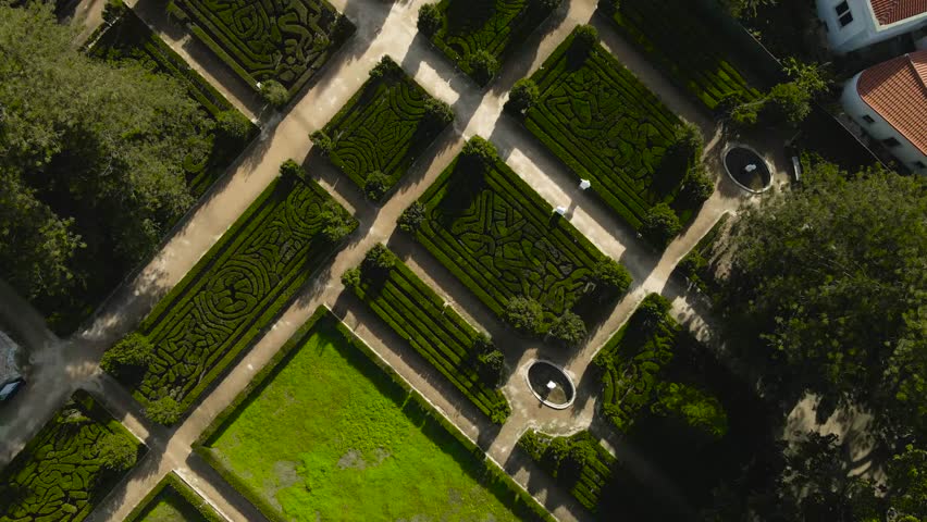 Top down aerial view of ornate baroque gardens with geometric hedges, fountains and pathways as tourists walk below on a sunny day.