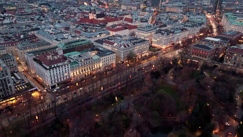Aerial high-angle landscape drone shot over Vienna cityscape near Stadtpark at night in winter, showing illuminated streets, city buildings, park outlines, glowing traffic, and winter night