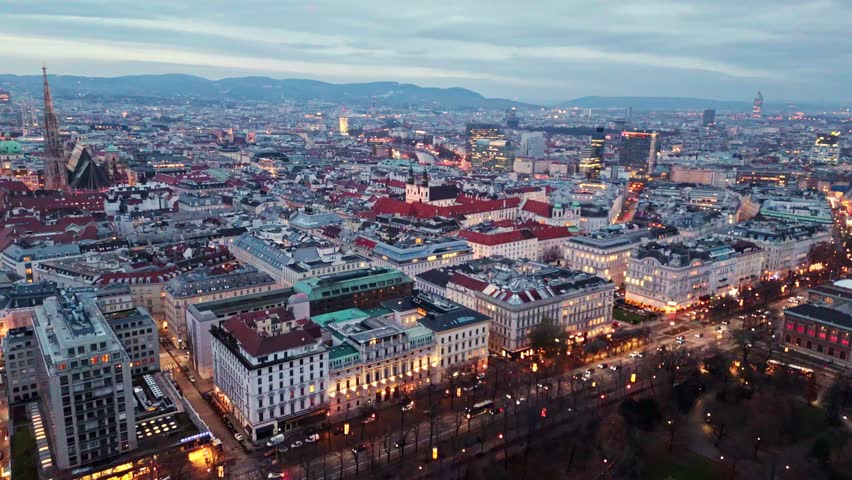 Aerial landscape static drone shot of Vienna cityscape at night in winter, showing illuminated buildings, glowing streets, flowing traffic, and city lights reflecting across rooftops under winter sky