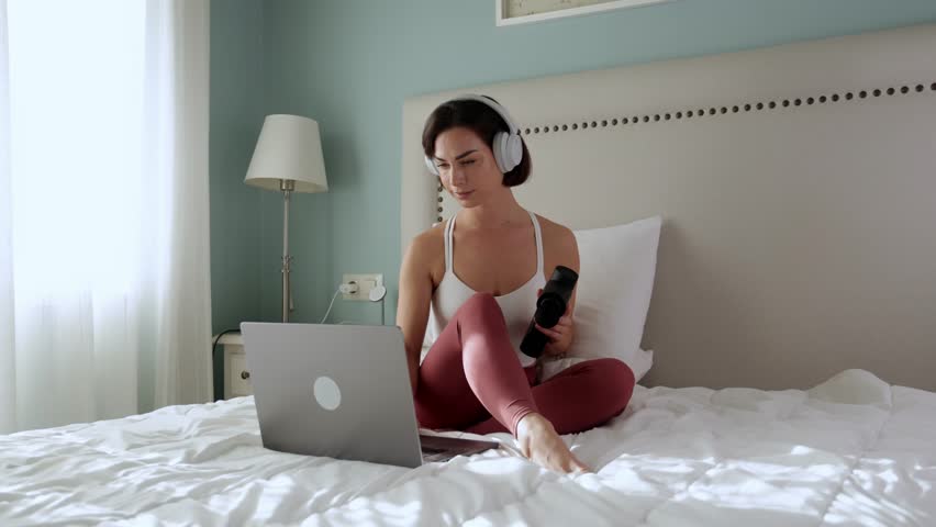 Sporty young woman using percussive massage gun at home while working on laptop. Physiotherapy, muscle recovery, self-massage tool. Daily lifestyle concept