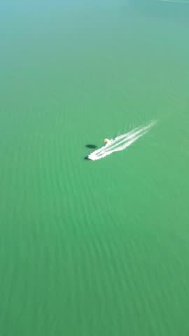 Vertical aerial parasailing over emerald sea. 4K drone footage shows a boat moving across vivid green water in Batumi, Georgia, creating white wake while towing a parachute in clear weather.