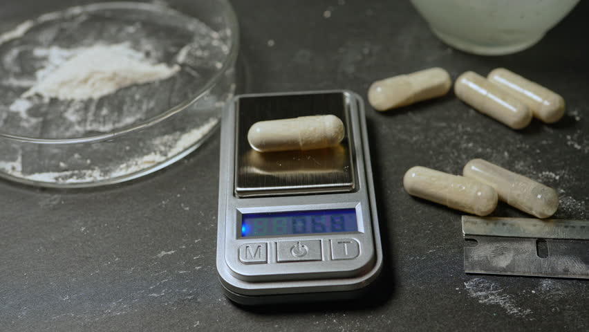 Drug dealer measure powder of synthetic opioid drug of fentanyl in the gelatin capsule for sale on the streets or parties, using electronic scale. Toxic drug crisis and struggle with an addiction.