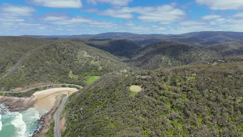 Aerial view of forested hills and coastal valley near the Great Ocean Road in Australia. Scenic landscape with winding road, green vegetation, mountains and ocean bay under a blue sky.
