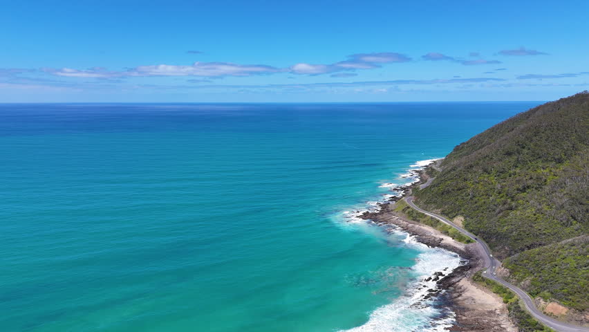 Aerial view of the Great Ocean Road winding along a rugged coastline, with turquoise ocean, rocky shore and green hills. Scenic coastal landscape and iconic seaside road in Australia.