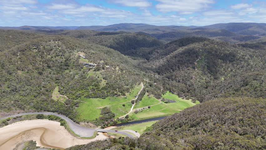Aerial view of forested hills and coastal valley near the Great Ocean Road in Australia. Scenic landscape with winding road, green vegetation, mountains and ocean bay under a blue sky.
