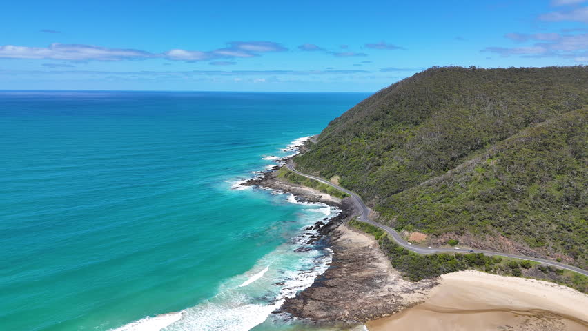 Aerial view of the Great Ocean Road winding along a rugged coastline, with turquoise ocean, rocky shore and green hills. Scenic coastal landscape and iconic seaside road in Australia.