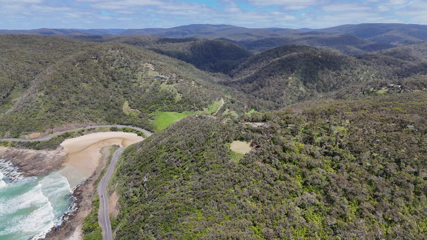 Aerial view of forested hills and coastal valley near the Great Ocean Road in Australia. Scenic landscape with winding road, green vegetation, mountains and ocean bay under a blue sky.
