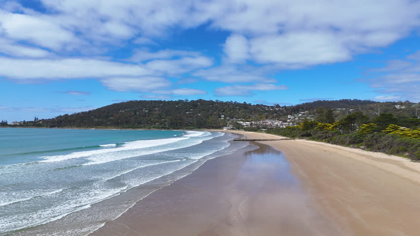 Aerial view of a curved sandy beach with gentle waves, turquoise sea and coastal town along green hills. Calm shoreline, natural coastline and seaside landscape on a clear day.