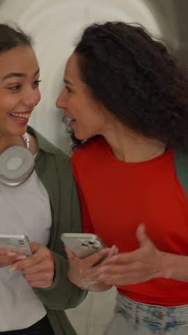 Two Hispanic girls students walk university corridor looking mobile phones laugh happy friends women Latina females walking going in high school college academy hall smiling talking gossip online app