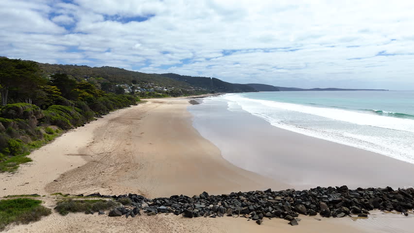 Wide coastal view of a long sandy beach with gentle waves, turquoise sea and forested hills. Scenic shoreline and seaside town under soft natural daylight.
