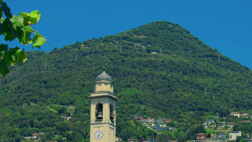 Historic clock tower rising above green hills near Lake Como under clear blue sky.