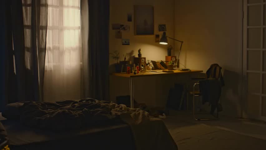 Pan shot of an inhabited bedroom in brown tones with an unmade bed and switched-on desk lamp at dawn, evoking insomnia and early morning mood.