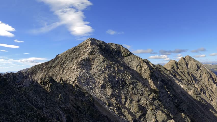Epic wide-angle drone footage looking down from a rugged rocky mountain peak, revealing a vast landscape of alpine lakes, deep valleys, and distant ranges under a blue sky.