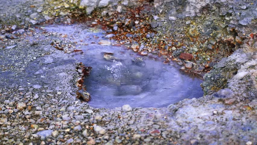 A detailed ground-level shot capturing a small geothermal vent bubbling with mineral-rich blue water and steam rising from the orange volcanic rim.
