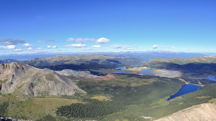POV style aerial shot looking down a sharp, steep mountain ridge with a vast valley and blue reservoirs visible in the distance under a clear sunny sky.