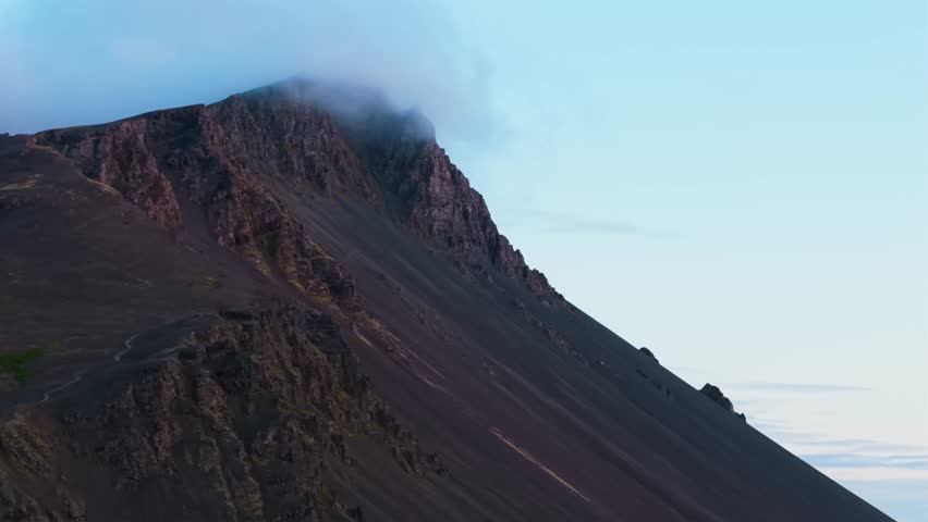 Moody and textured drone shot focusing on the steep, loose scree slopes and jagged rock pinnacles of a dark volcanic mountain in Iceland under cloudy skies.