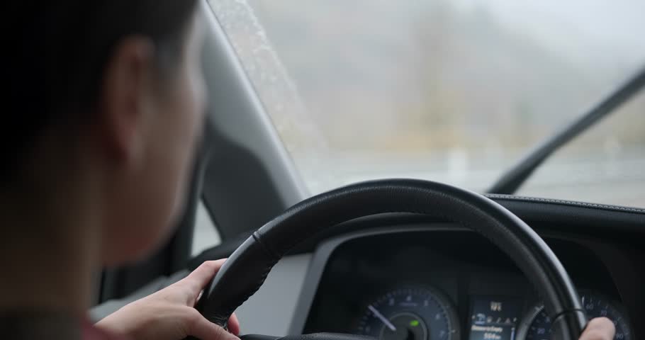 Over the shoulder view of a woman driving a car during heavy rain. Hands on steering wheel, windshield wipers clearing water from the glass. Bad weather driving.