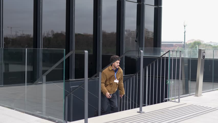 Young male employee with an id badge frantically running up the stairs outside a modern office building, holding a phone with a look of urgency