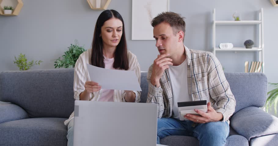 Young family couple sitting on sofa in front of laptop with paper sheet and calculator and discussing variants. Husband and wife counting cost of different alternatives and making decision together.