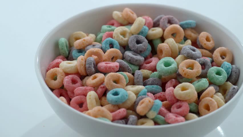 A high-quality close-up shot of fresh white milk being poured into a white ceramic bowl filled with vibrant, multi-colored ring-shaped cereal. The milk splashes gently, causing the cereal to float and shift, with a few pieces spilling over the edge. Set against a clean white background, this bright and energetic scene captures a classic morning breakfast routine.