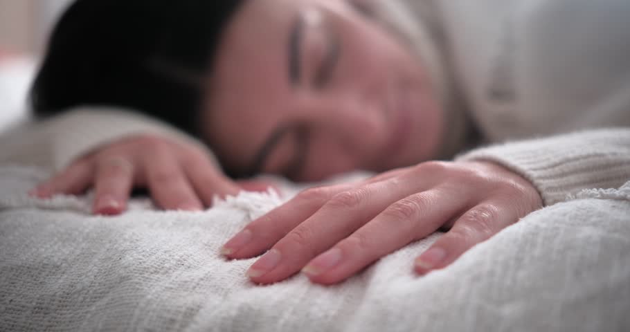 Close up of female hand gently touching soft white texture of blanket on bed. Blurred smiling woman sleeping or waking up in background. Cozy morning atmosphere.