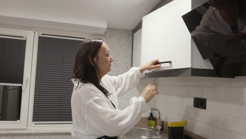 Middle aged caucasian woman taking white plates from kitchen cabinet indoors, everyday dish organization in modern kitchen interior.