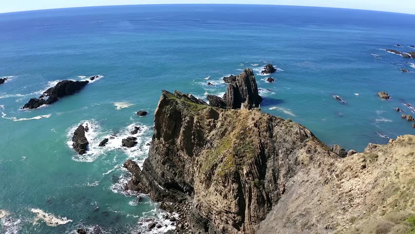 Spectacular aerial drone shot orbiting a sharp, jagged rock formation jutting out of the deep blue ocean. The sunlight highlights the rugged texture of the cliffs against the vibrant water.