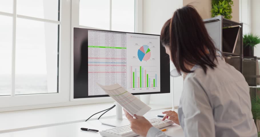 Analyst woman reviews data charts on computer monitor. She reviews an office dashboard and spreadsheet, interprets metrics, and prepares a report for planning. Clear data driven decision concept.