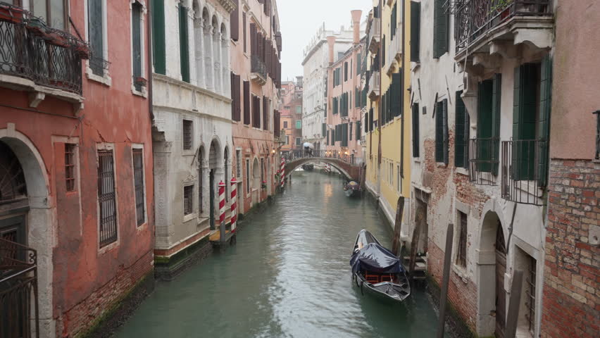 White motorboat navigating narrow Venice canal between historic buildings during rain. Water taxi moving toward camera with moored gondola and people on bridge under umbrellas in Italy.