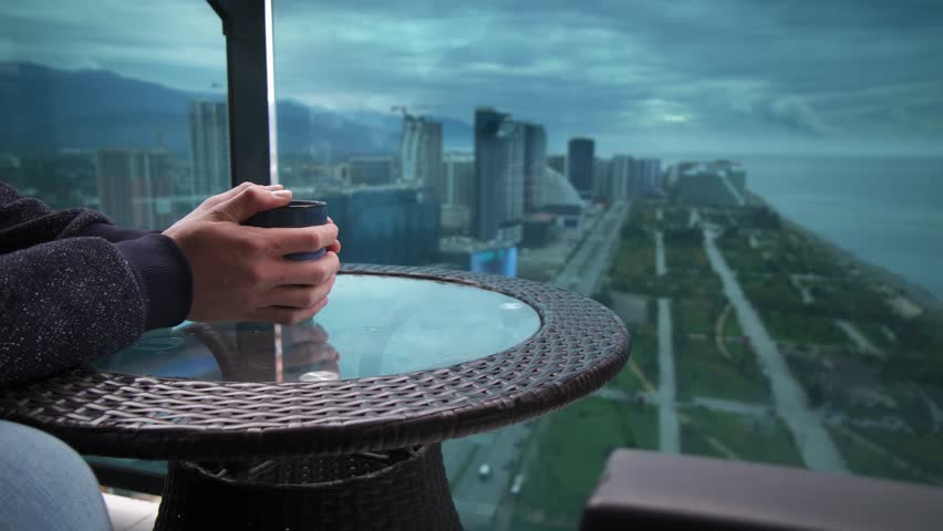 man drinking tea on balcony Batumi skyline and Black Sea view at dusk, hands holding ceramic cup on wicker table, distant promenade and highrises fading into mist, solitary traveler reflecting