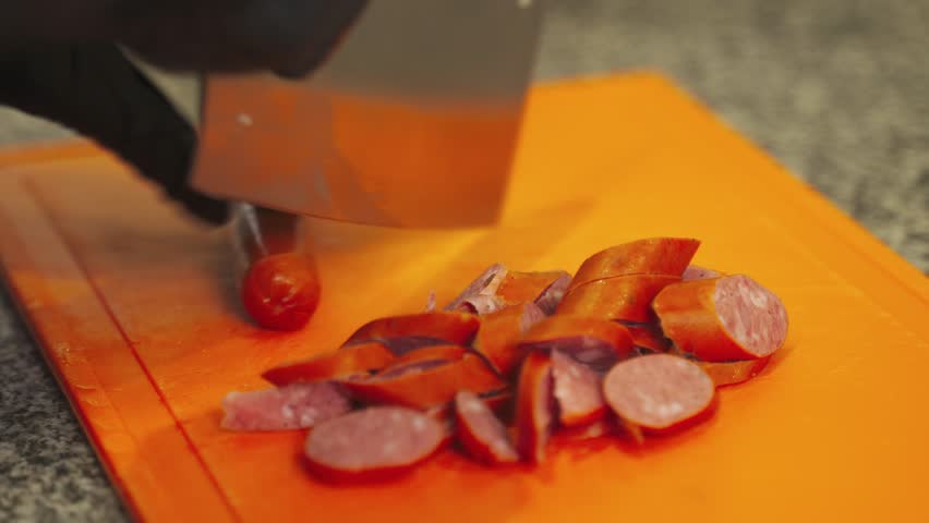 Close-up view of succulent grilled sausage slices being expertly chopped on a vibrant cutting board. The aroma fills the air as this savory treat is prepared. Ideal for your next meal.