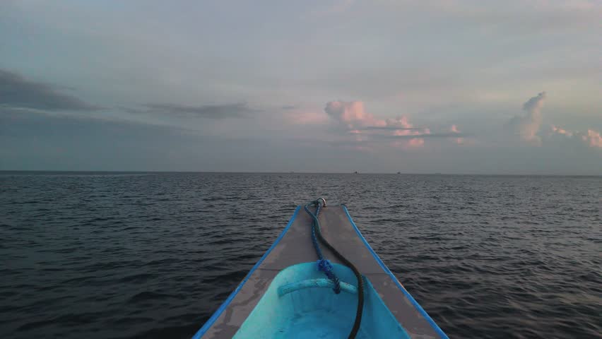 POV 4K footage from a blue traditional boat gliding toward the sunrise horizon over tranquil ocean waters. Warm golden rays illuminate scattered clouds and gentle waves. 
