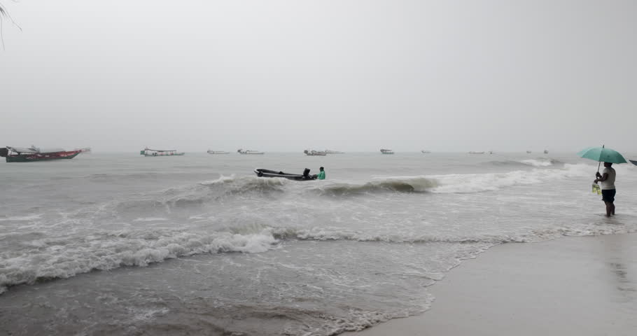 Man in a raincoat struggling with a small boat in rough waves during a storm, with a person holding an umbrella on the beach,  with several fishing boats in the background.