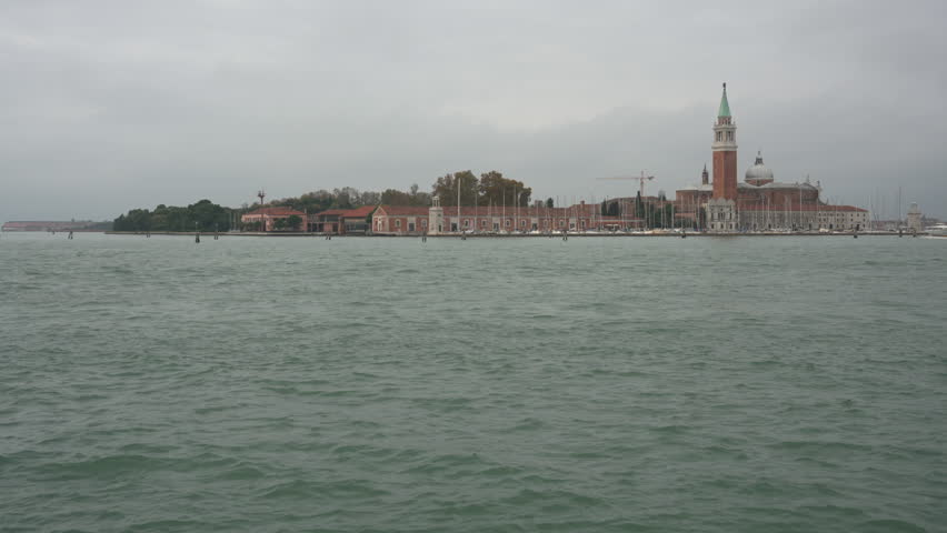 View of San Giorgio Maggiore island across Venetian lagoon under grey overcast sky. Historic Renaissance church and bell tower landmark. Moody atmospheric Italy travel landscape.