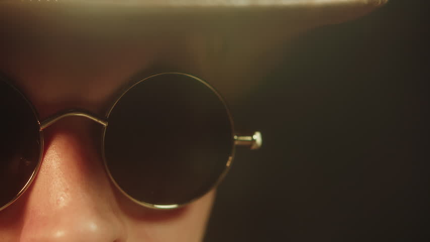 A close-up of a young adult caucasian man wearing dark glasses and a cowboy hat, looking at the camera and speaking. A portrait shot of an actor performing a role against a dark studio background.